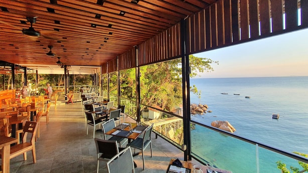 Un espace de restauration en plein air, avec des tables et des chaises, offrant une vue sur un plan d’eau et une côte rocheuse.