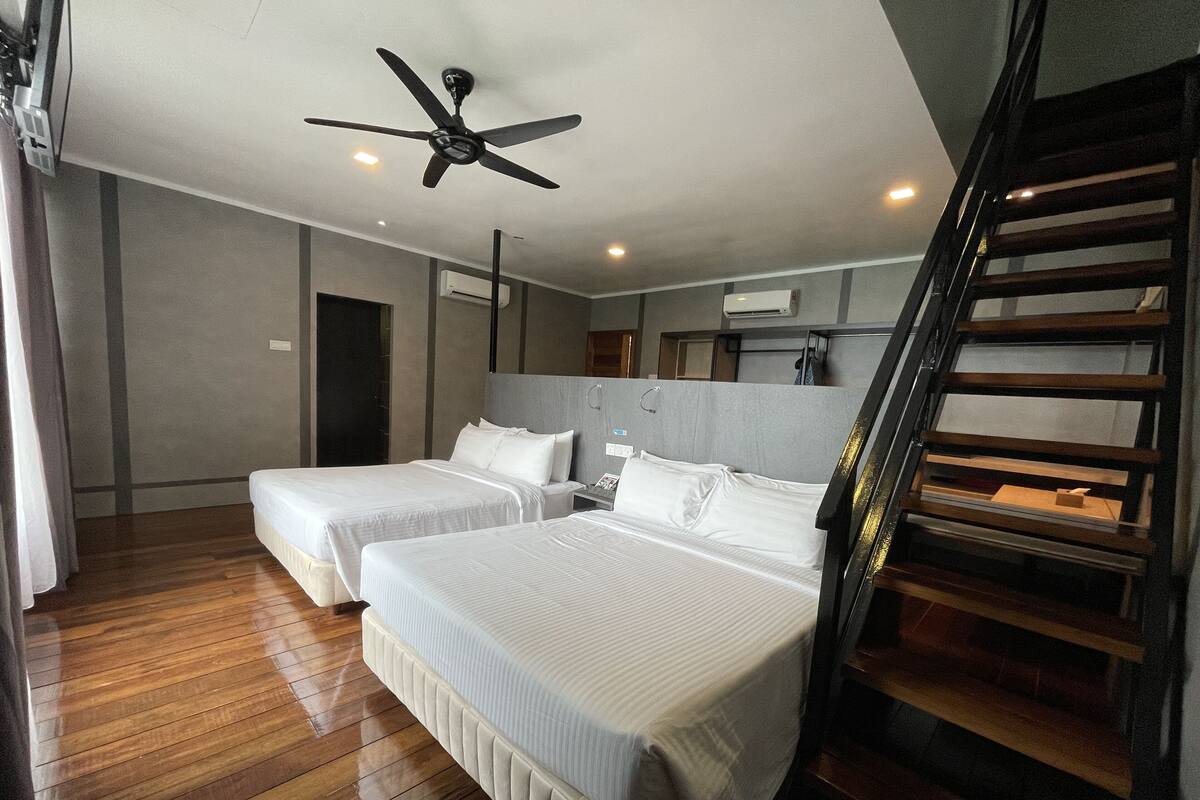 A hotel room with two beds, a ceiling fan, wooden flooring, a staircase, and a view of the outside.