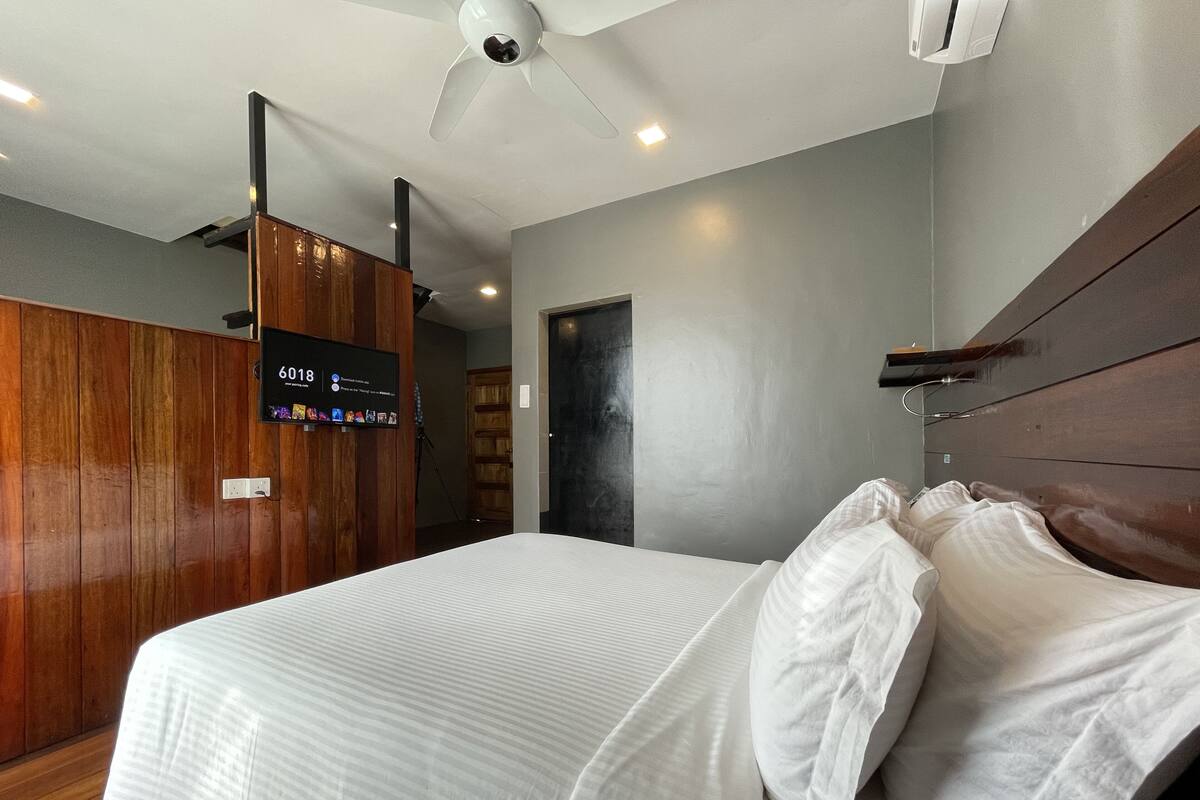 A modern hotel room with a large bed, wooden paneling on one wall, a ceiling fan, and a mounted TV displaying channel 6018.