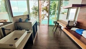 Une salle de bain moderne avec une baignoire, un lavabo et un espace douche. On y trouve une chaise et une petite table avec une machine à café. La pièce est dotée de grandes fenêtres offrant une vue sur l’océan et les arbres.