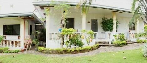 A two-story house with a porch, surrounded by lush greenery and palm trees.
