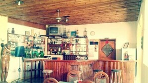 Un coin bar avec un plafond en bois, un téléviseur et diverses bouteilles. On y trouve des chaises et une table recouverte d’une nappe colorée.