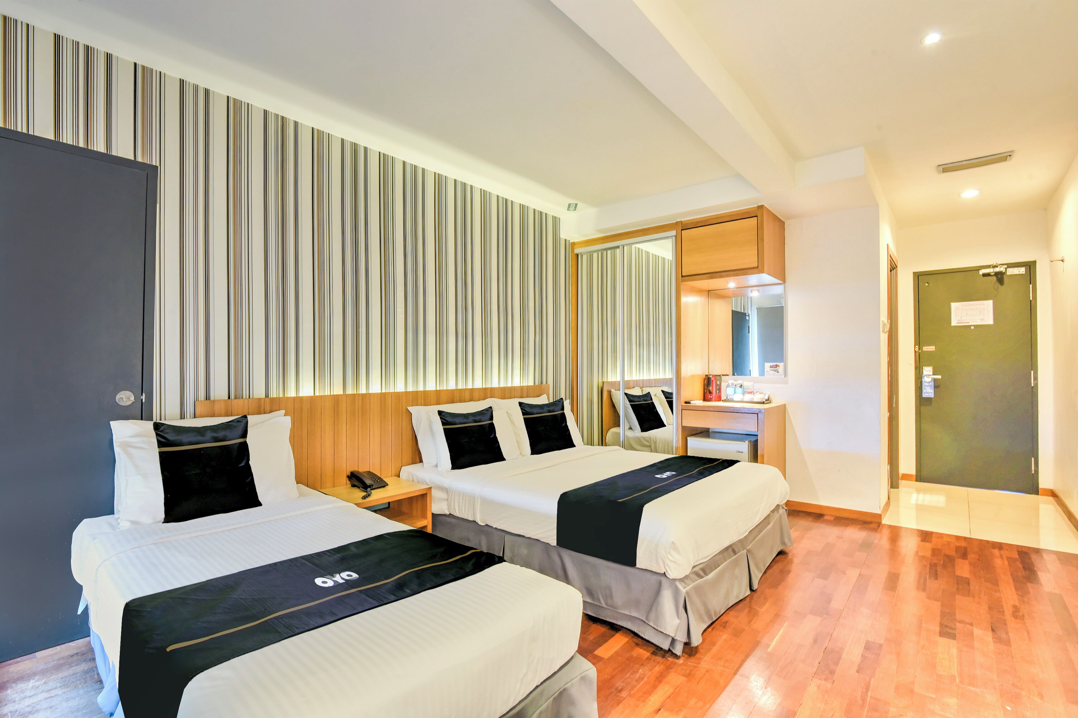 A hotel room with two beds, a wooden headboard, a striped wall, a mirror, a desk with a phone, and a green door.
