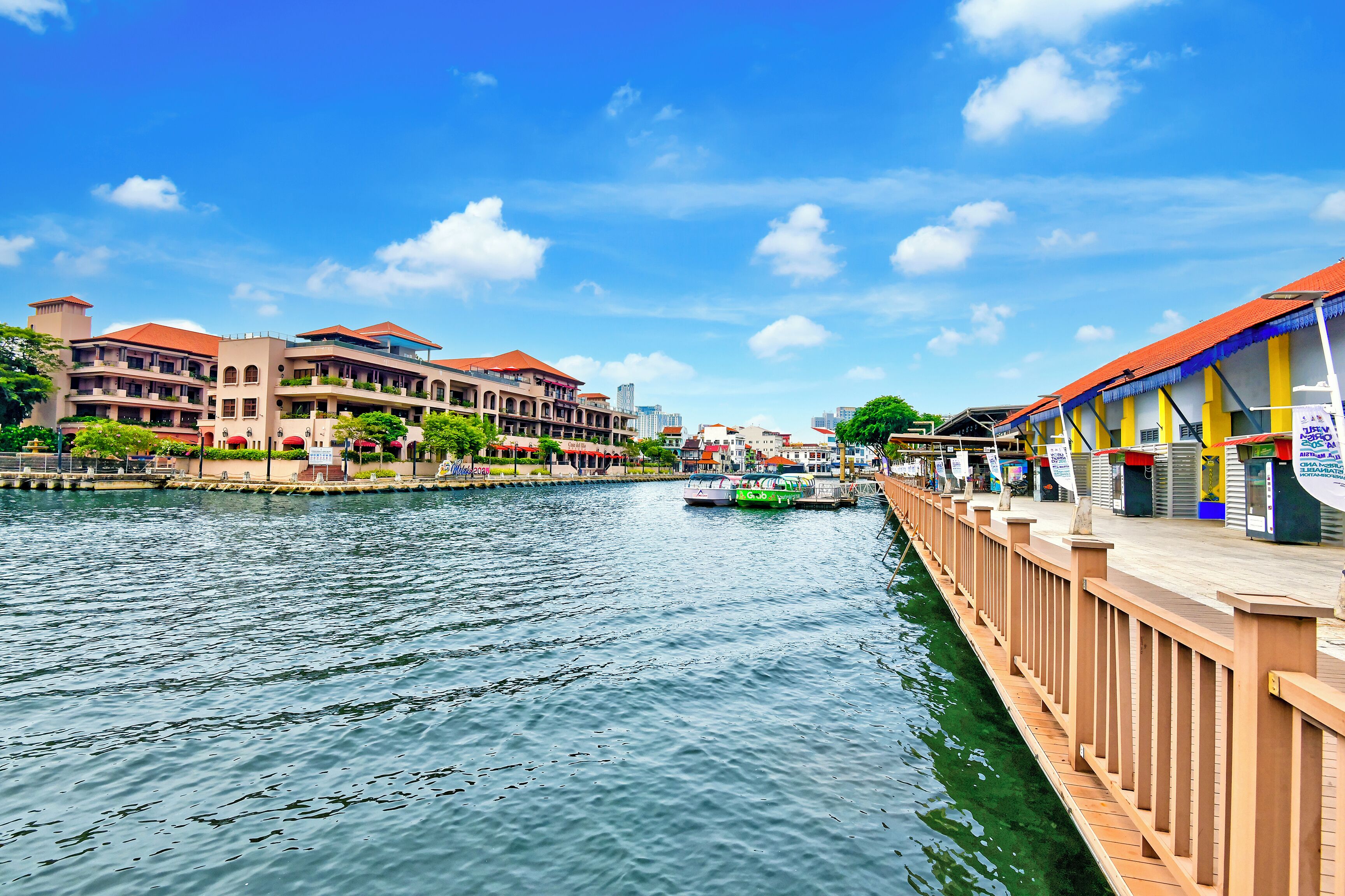 A waterfront hotel with multiple balconies, a river in the foreground, and a bridge connecting to the opposite bank.