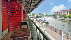 A wooden deck with a red wall and a metal railing overlooking a river with boats and buildings in the background. - Sunday Malacca Near Jonker Street Formerly Quayside (Malacca City)