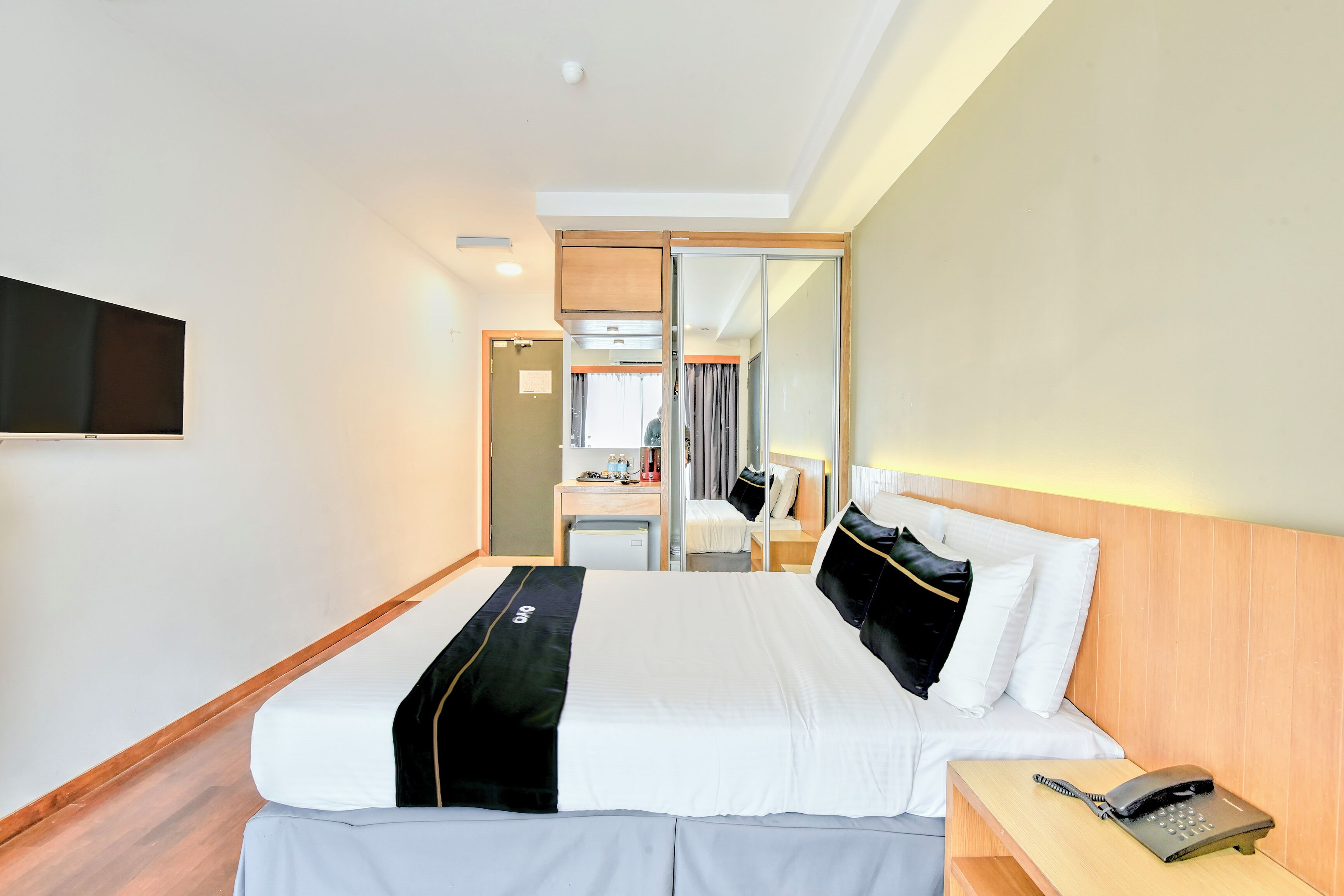 A hotel room with a bed, bedside table with a phone, a desk with a chair, a mirror, and a TV mounted on the wall.