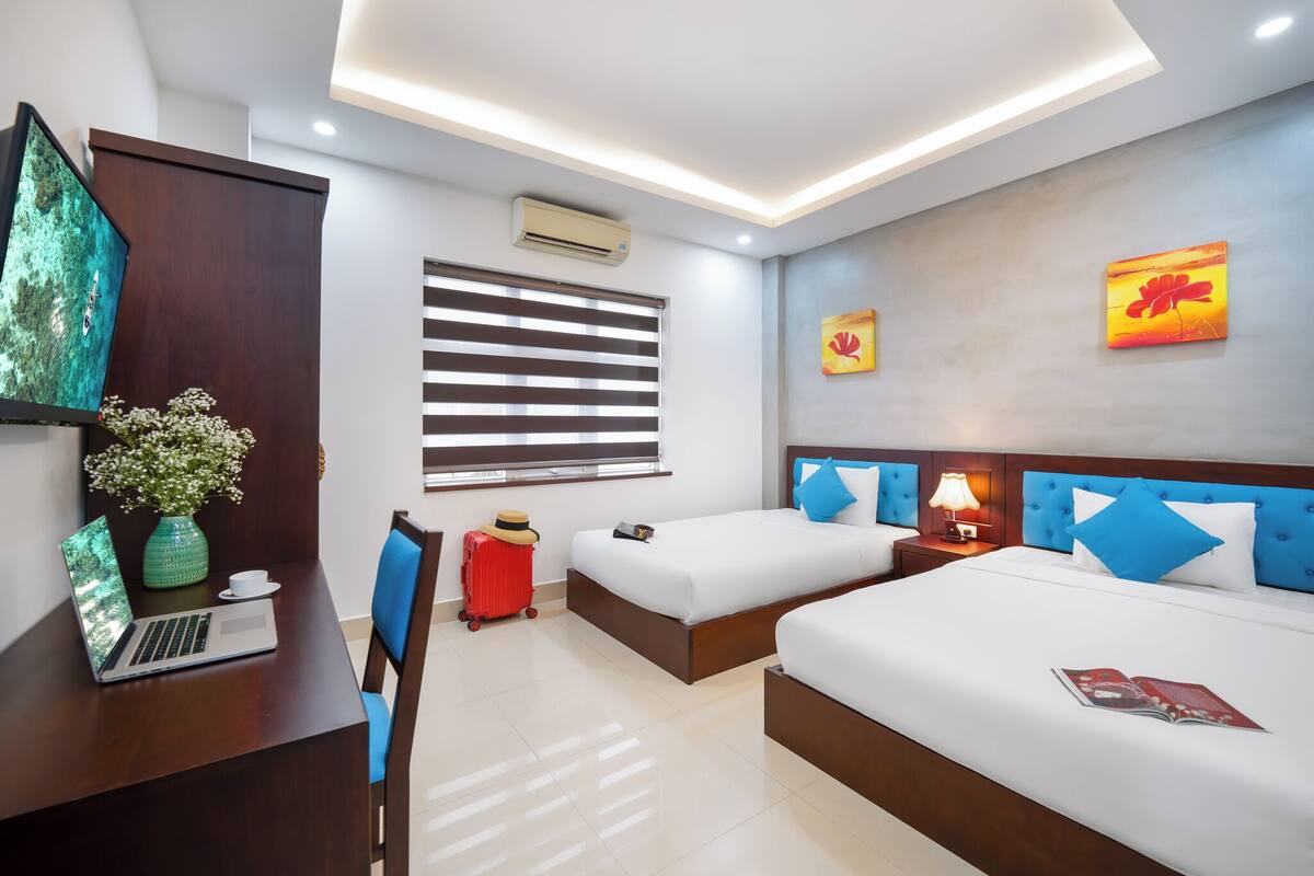 Habitación de hotel con dos camas, un escritorio con una computadora portátil, un florero con flores, una maleta y una ventana con persianas.