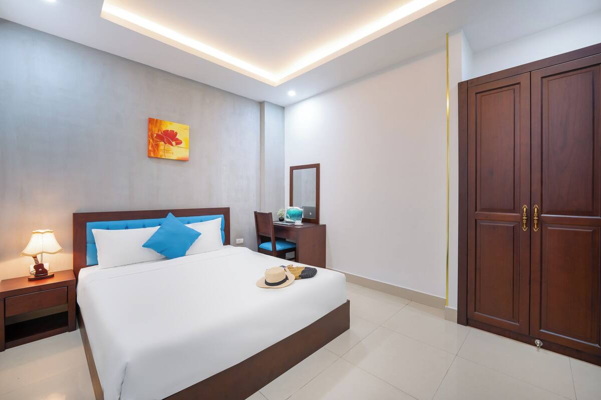 Una habitación de hotel moderna con una cama grande, una mesita de noche con lámpara, un escritorio con silla, un armario y una obra de arte en la pared.