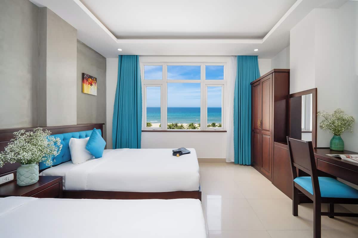 Habitación de hotel con dos camas, vista al océano, un escritorio con silla y un armario.