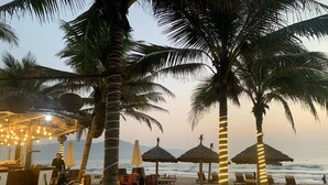 A beachside bar with thatched umbrellas, palm trees, and a wooden deck. - Platinum Beach Hotel (Da Nang)