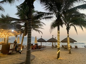 A beachside bar with thatched umbrellas, palm trees, and a wooden deck. - Platinum Beach Hotel (Da Nang)
