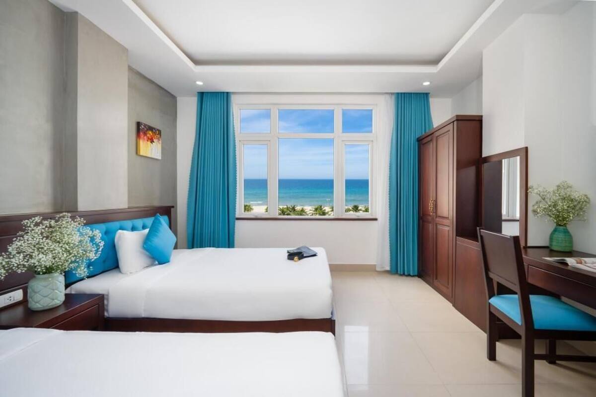 Habitación de hotel con dos camas, un escritorio, una silla, un armario y un amplio ventanal con vistas al océano.