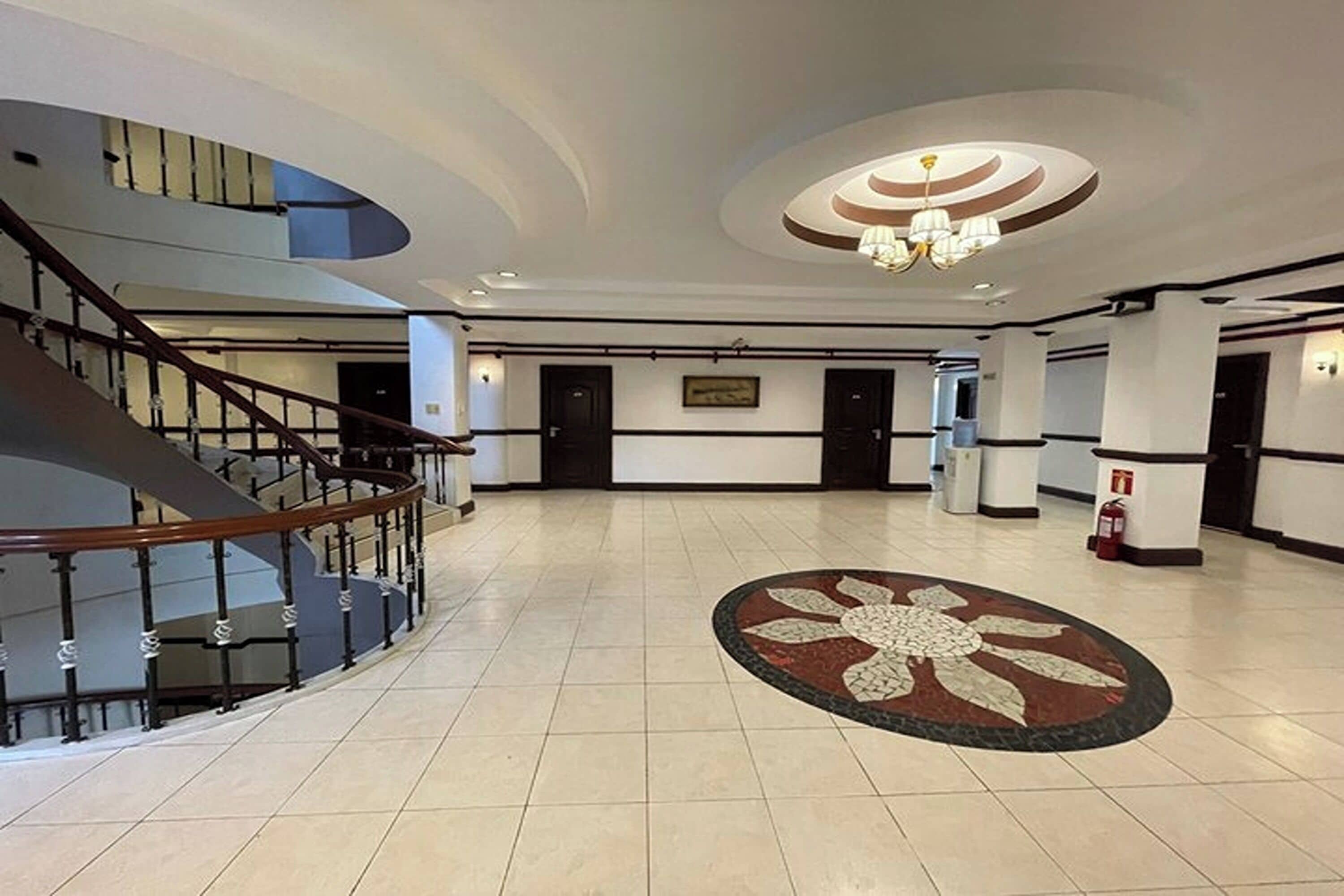 A spacious lobby with a central floral carpet, a curved staircase, and a chandelier.