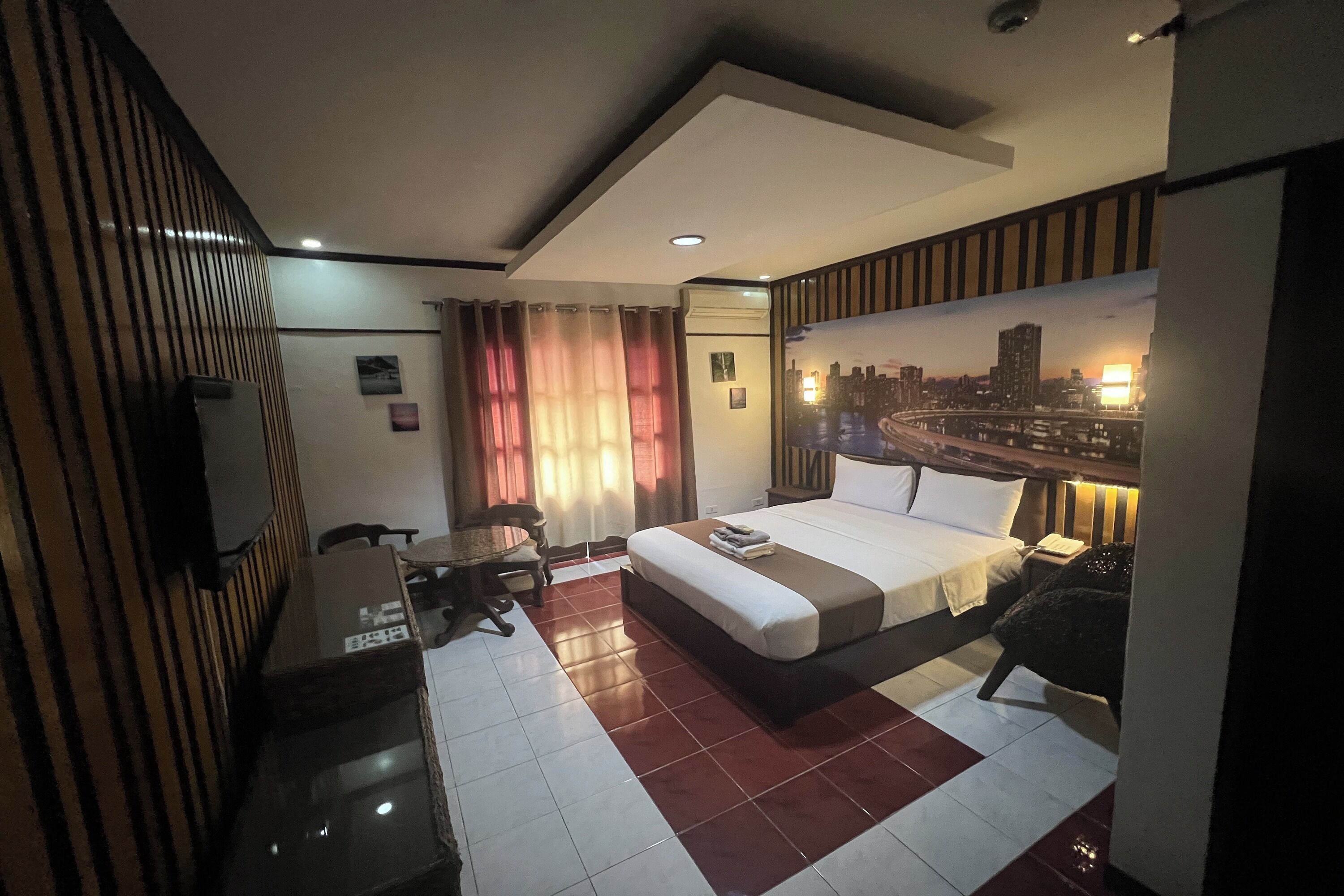 A hotel room with a bed, a desk, a chair, a small table, and a cityscape mural on the wall.