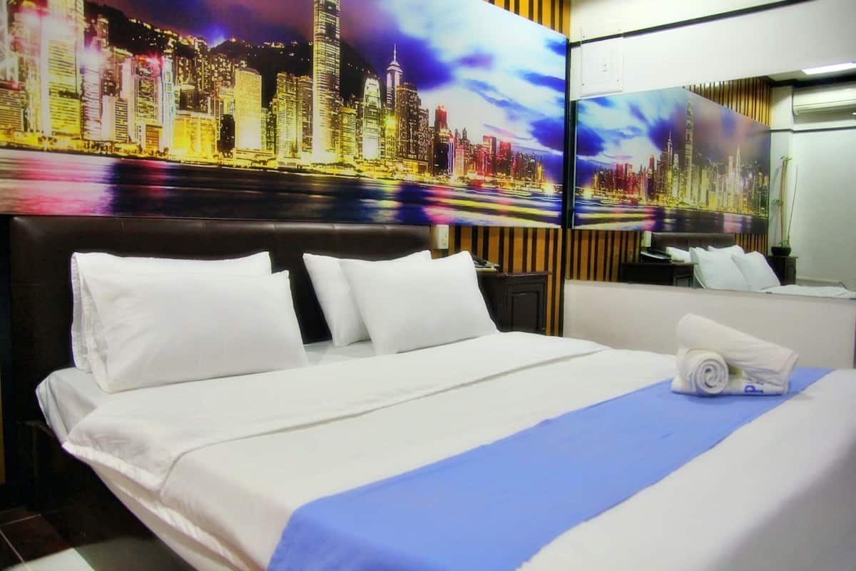 A hotel room with a bed, white bedding, a blue accent on the bedspread, a cityscape mural on the wall, and a night sky view.