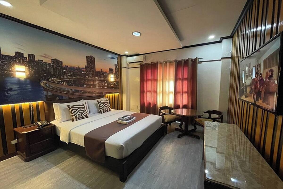 A hotel room with a bed, bedside table, nightstand, desk, chair, and a cityscape mural on the wall.