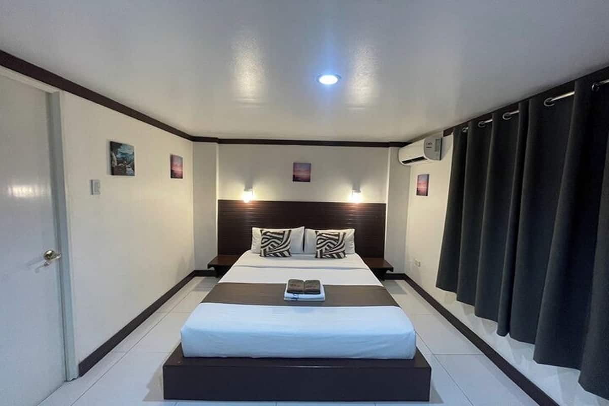 A hotel room with a large bed, a bedside table, a nightstand, a window with curtains, and a ceiling light.