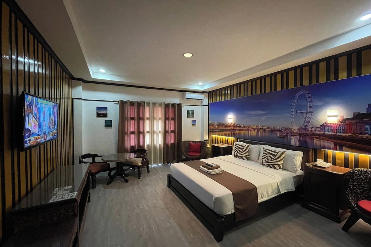 A hotel room with a bed, a desk, a chair, a TV, and a large cityscape mural on the wall.