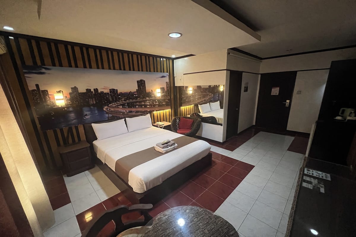 A hotel room with a bed, a small table, a chair, a nightstand, and a cityscape view on the wall.