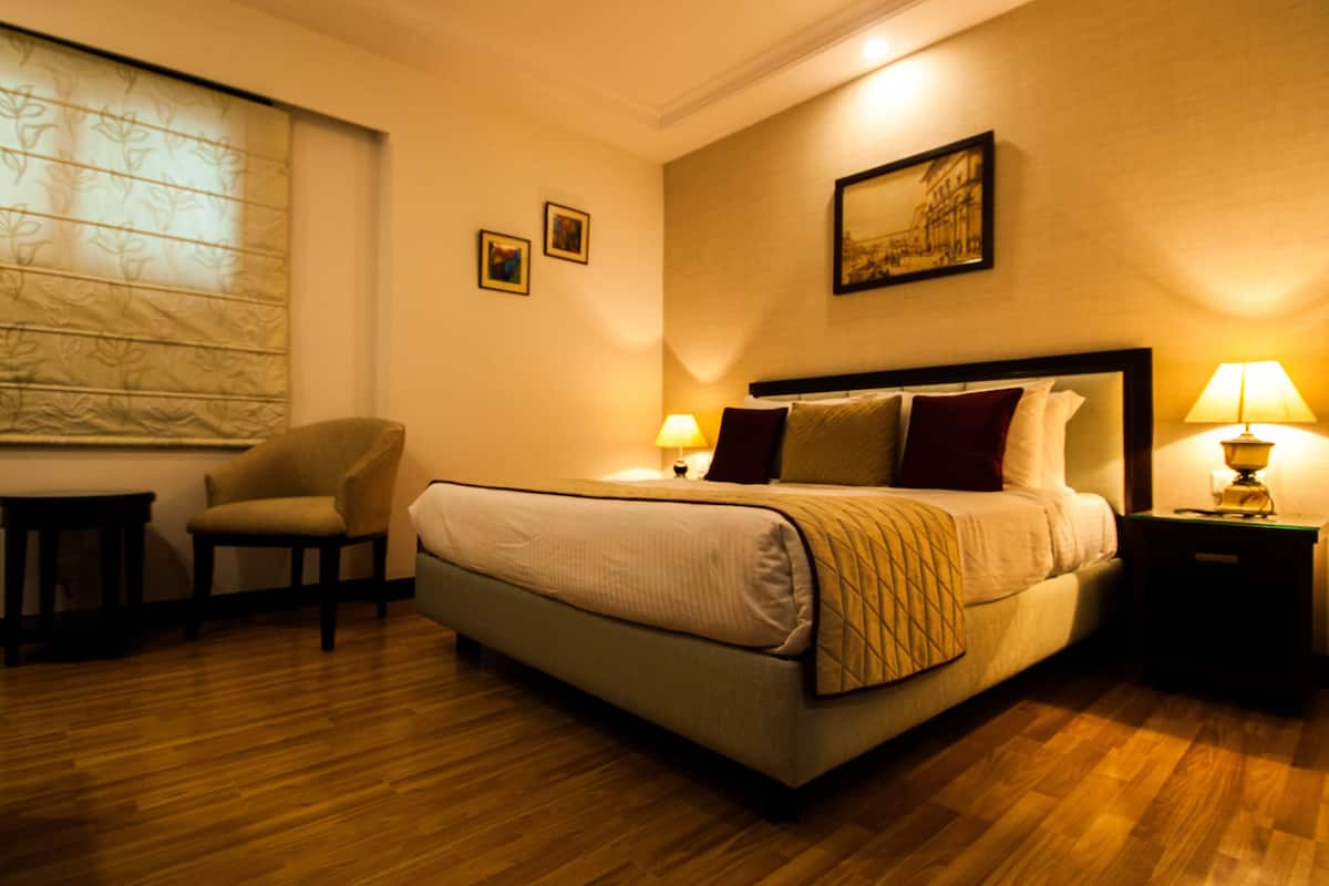 Una habitación de hotel con una cama, dos mesitas de noche con lámparas, una silla, una ventana con persianas y un cuadro en la pared.