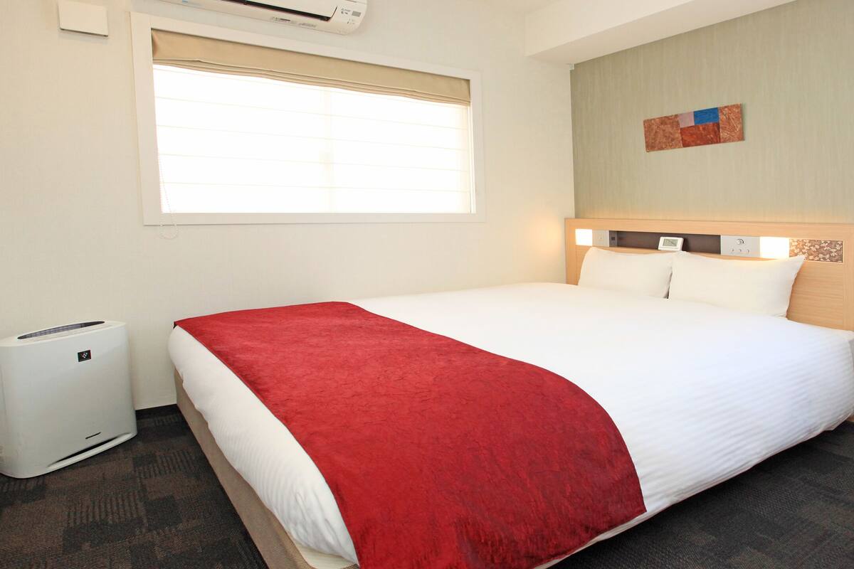 Une chambre d’hôtel comprenant un lit, une table de chevet, une fenêtre avec stores, un appareil de climatisation et une œuvre d’art accrochée au mur.