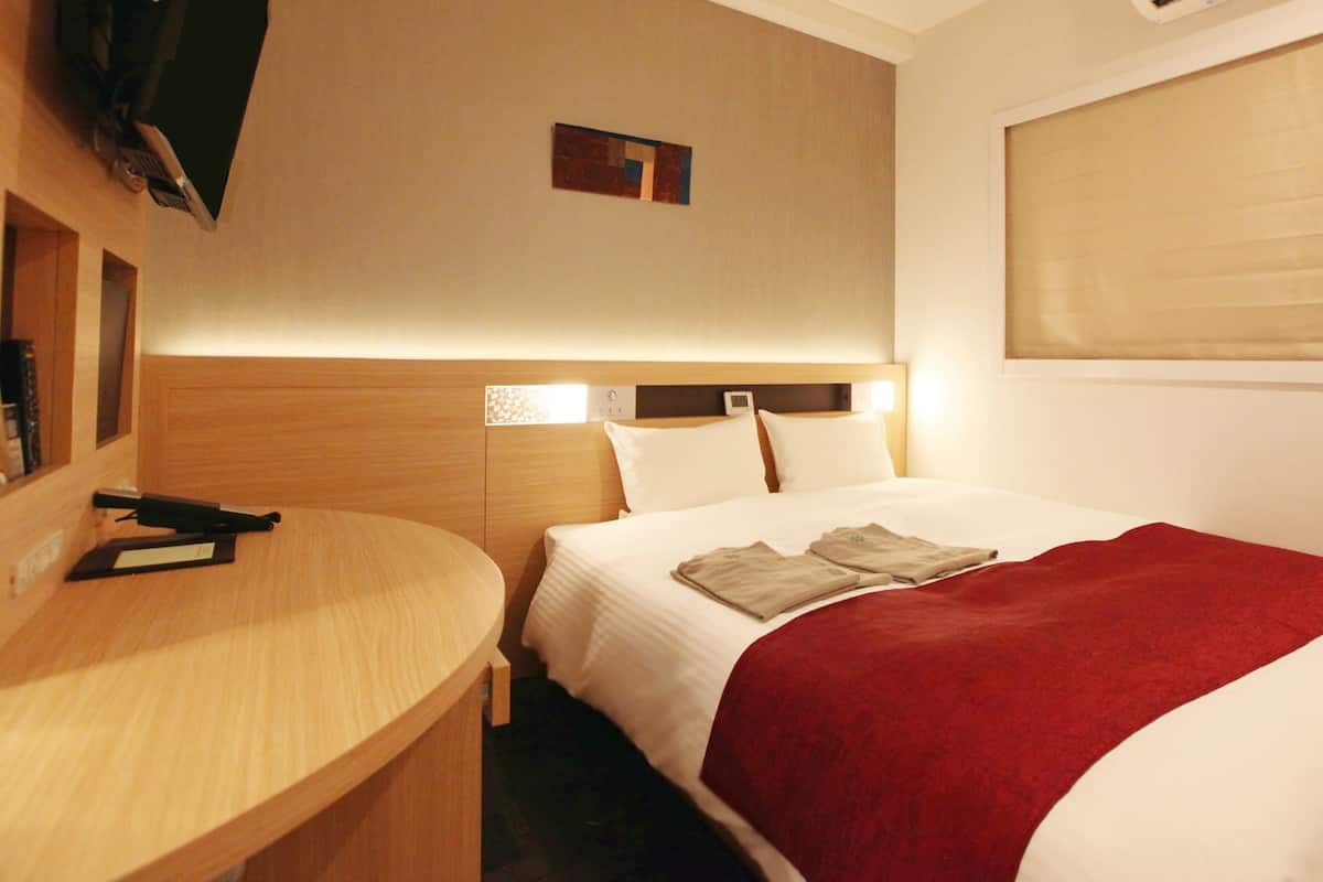 Une chambre d’hôtel avec un lit, un bureau, une télévision et une fenêtre avec des stores.