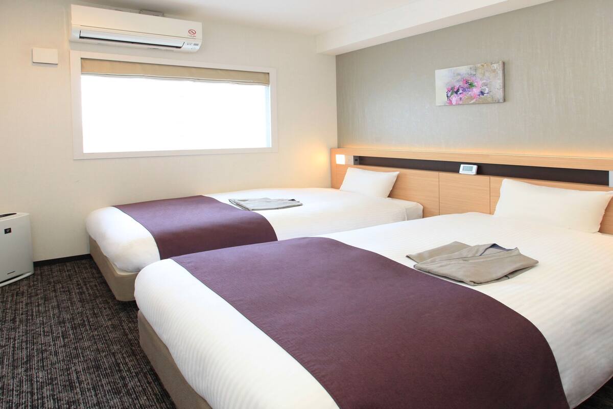 Une chambre d’hôtel avec deux lits, une fenêtre avec des stores, un climatiseur et un tableau au mur.