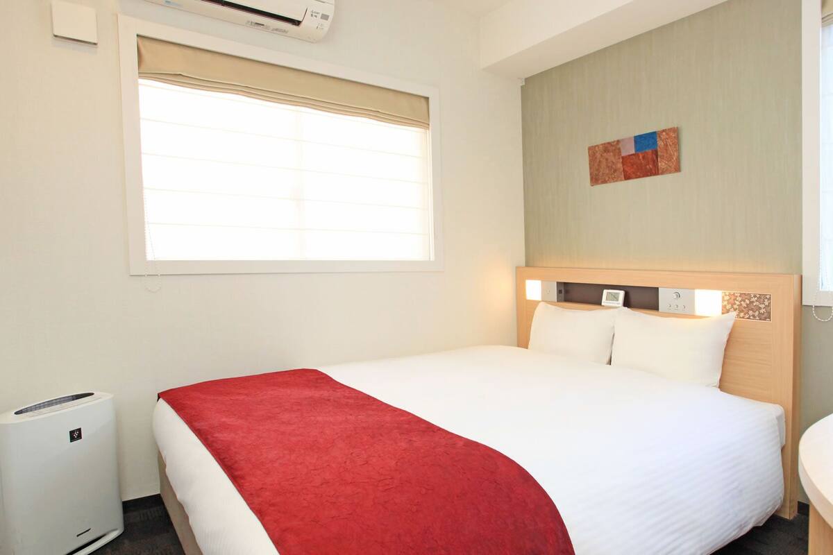 Une chambre d’hôtel comprenant un lit, une table de chevet, une fenêtre avec stores, un appareil de climatisation et une œuvre d’art accrochée au mur.