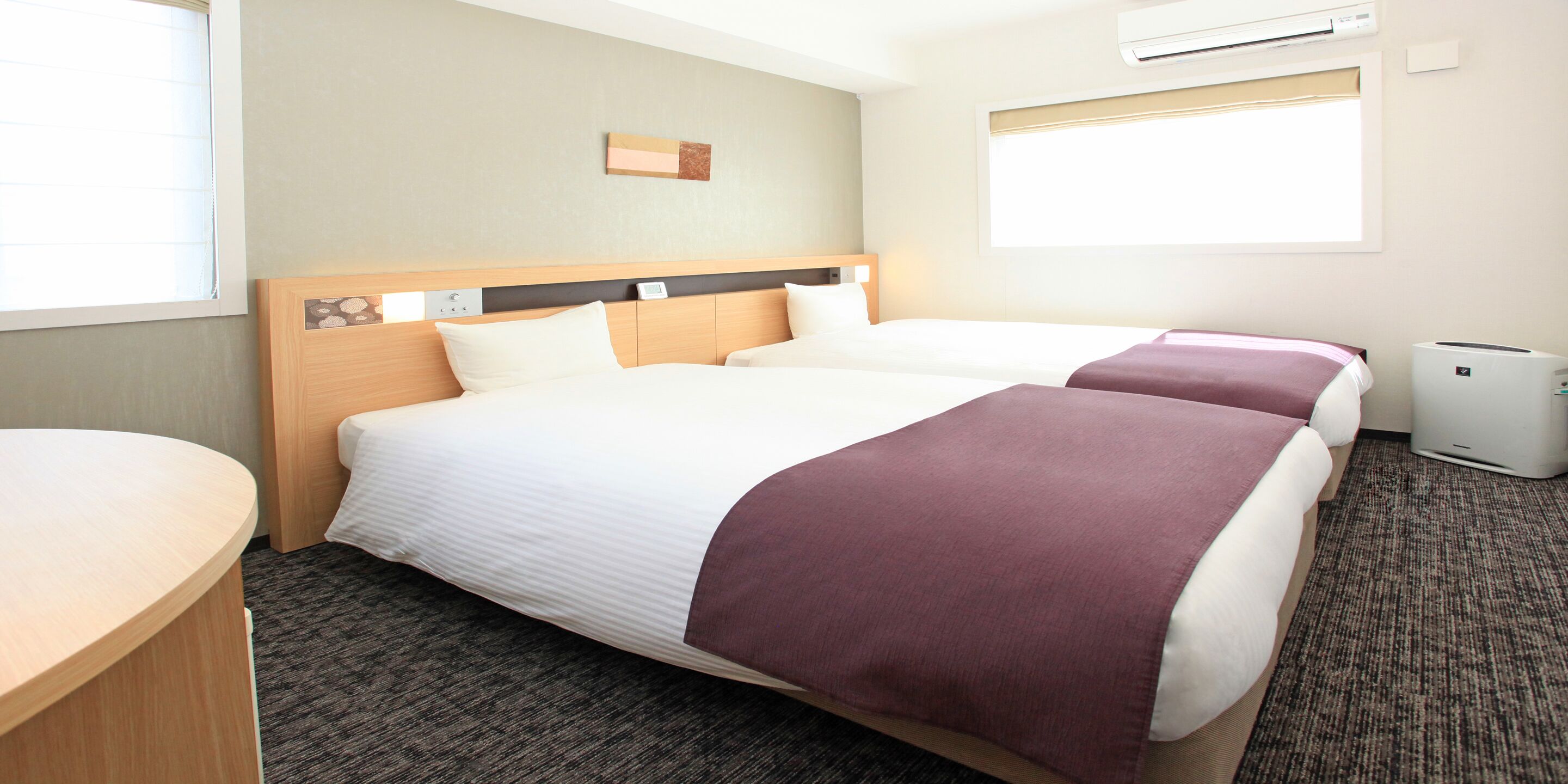 A hotel room with two beds, a wooden headboard, a desk, and an air conditioning unit.