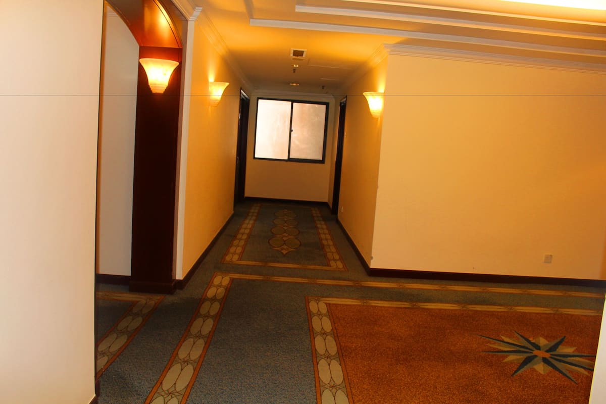 A hotel hallway with a carpeted floor, a window at the end, and wall-mounted lights.
