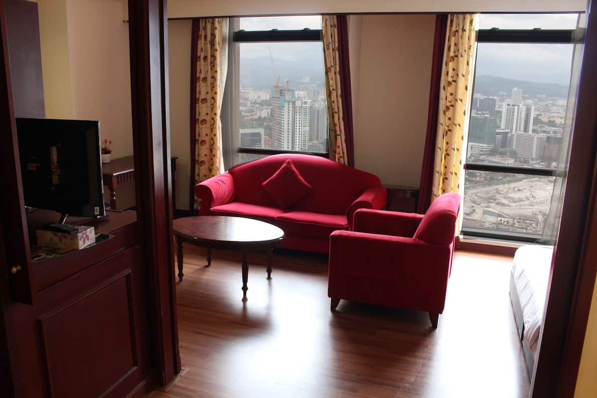 A hotel room with a red sofa, a small round table, and a TV on a wooden cabinet. A large window offers a view of the cityscape.