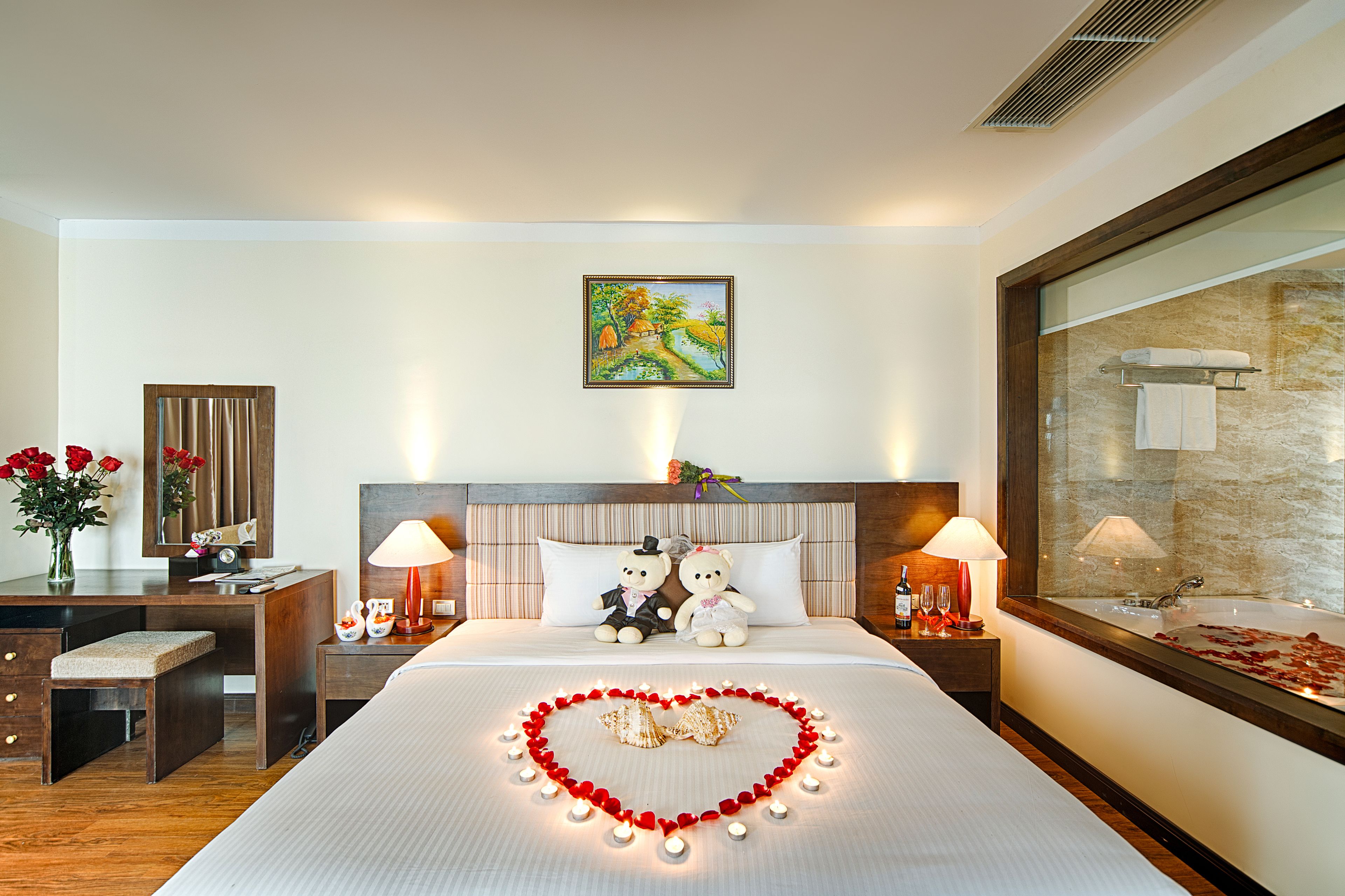 A hotel room with a bed, bedside lamps, a desk, a chair, a mirror, a painting, and a bathroom with a bathtub and towel rack.