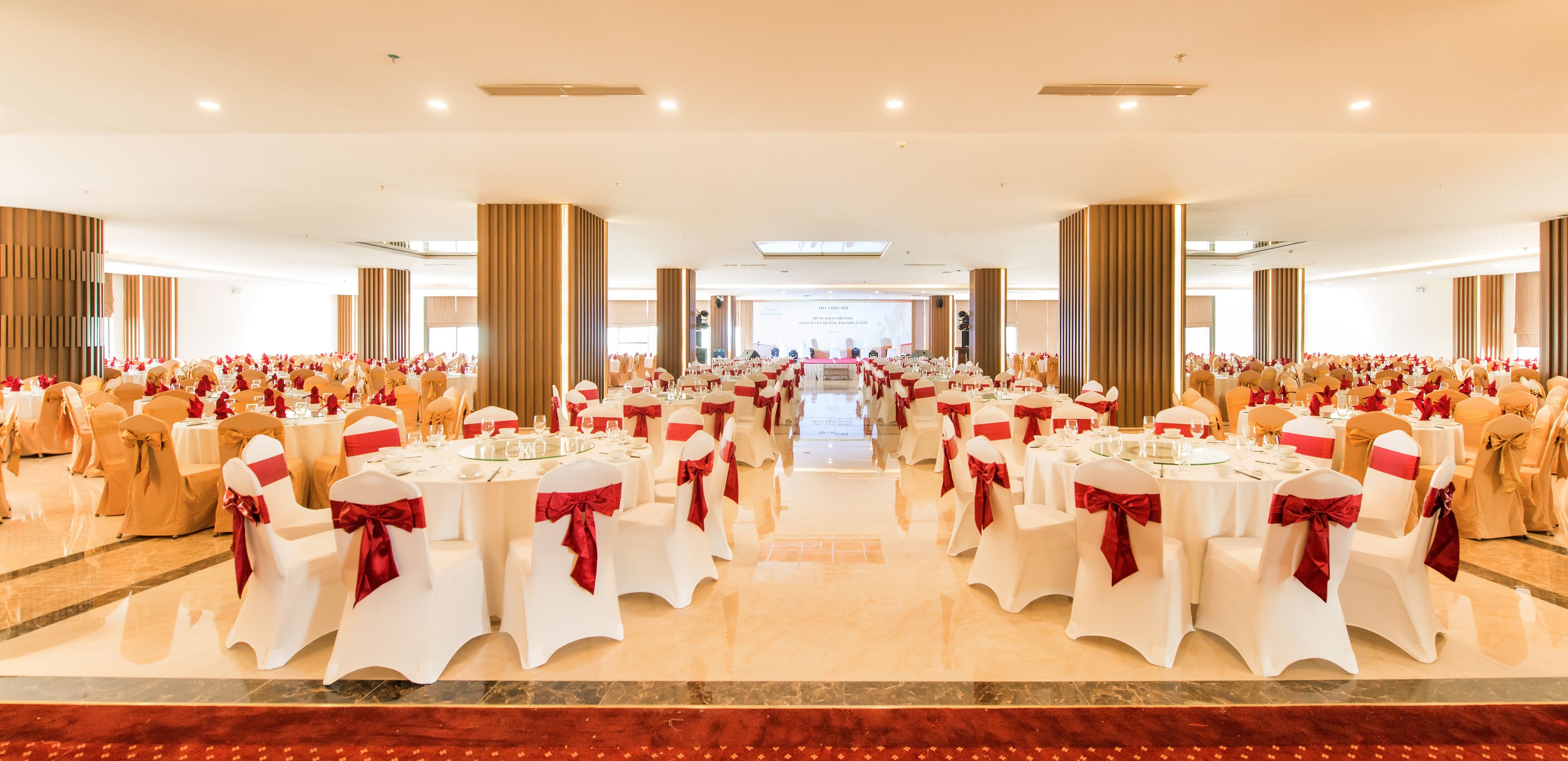 A banquet hall with round tables, white chairs with red sashes, and a stage at the front.