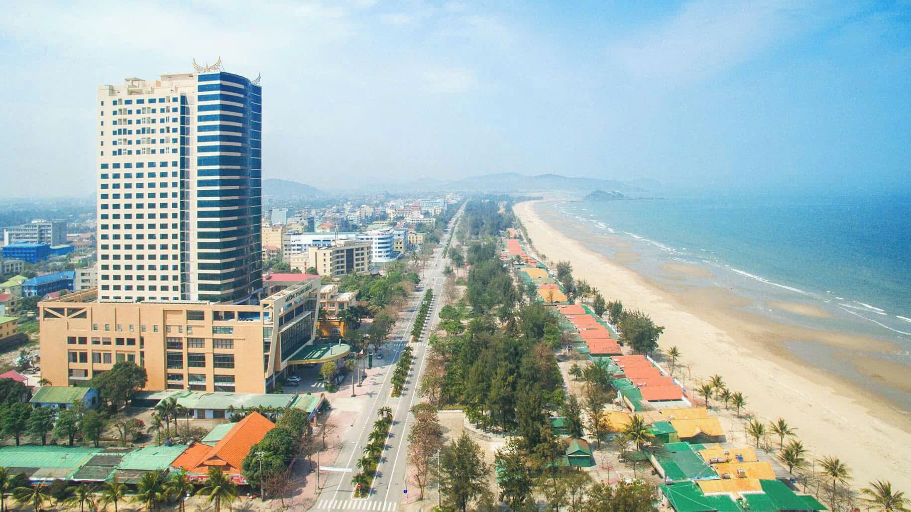 Un paysage urbain côtier avec un immeuble de grande hauteur qui domine le paysage, une plage et une route bordée de bâtiments aux couleurs vives.