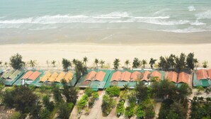 Un hotel frente a la playa con filas de habitaciones de techo verde, palmeras y el océano al fondo.