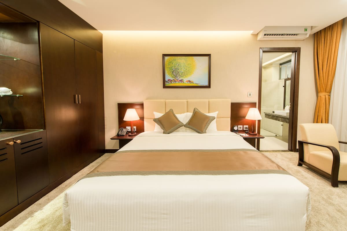 A hotel room with a large bed, two bedside tables with lamps, a nightstand, a wardrobe, a bathroom visible through an open door, and a painting on the wall.