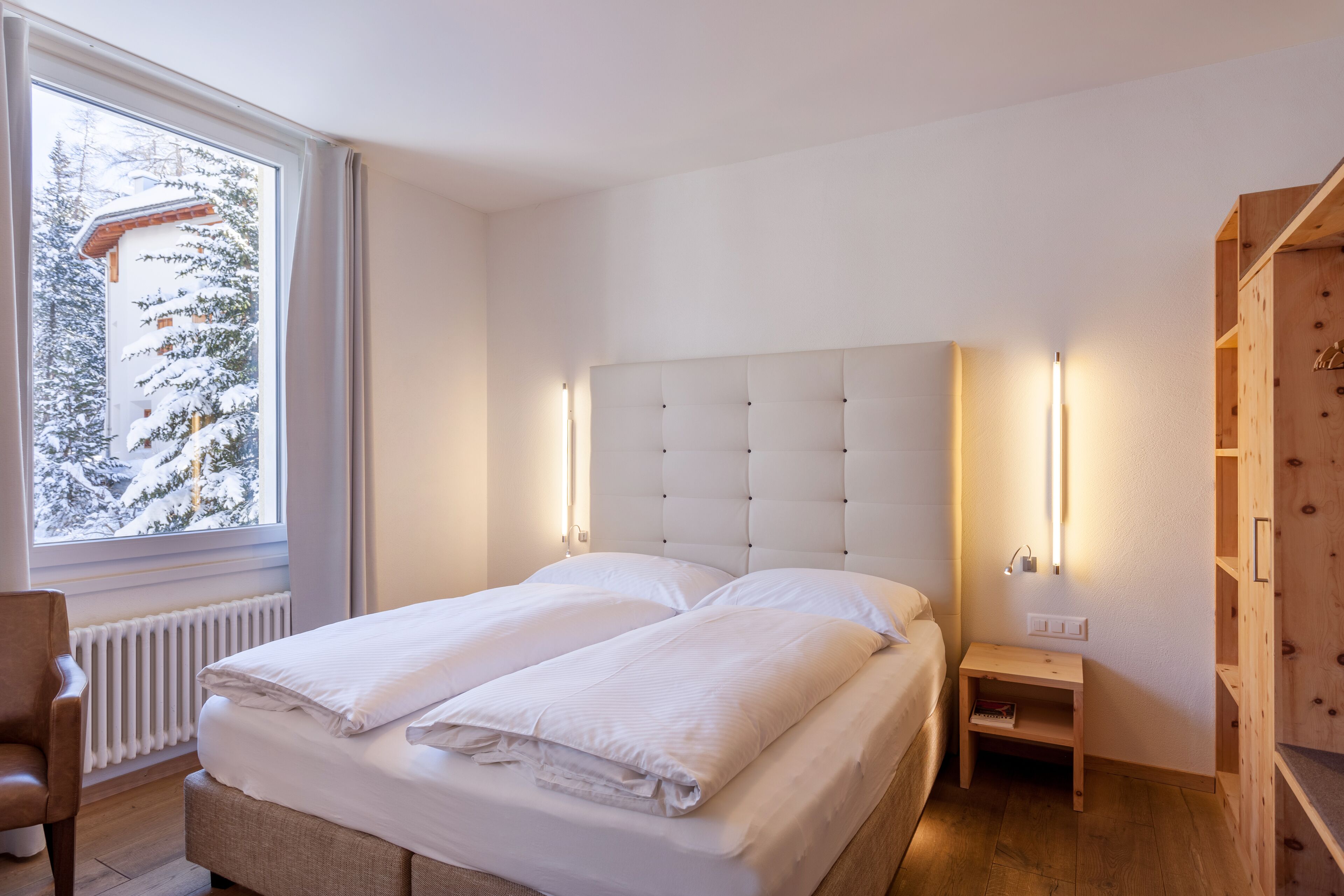 A bedroom with a large bed, a nightstand, a wooden wardrobe, and a window with a view of snow-covered trees.