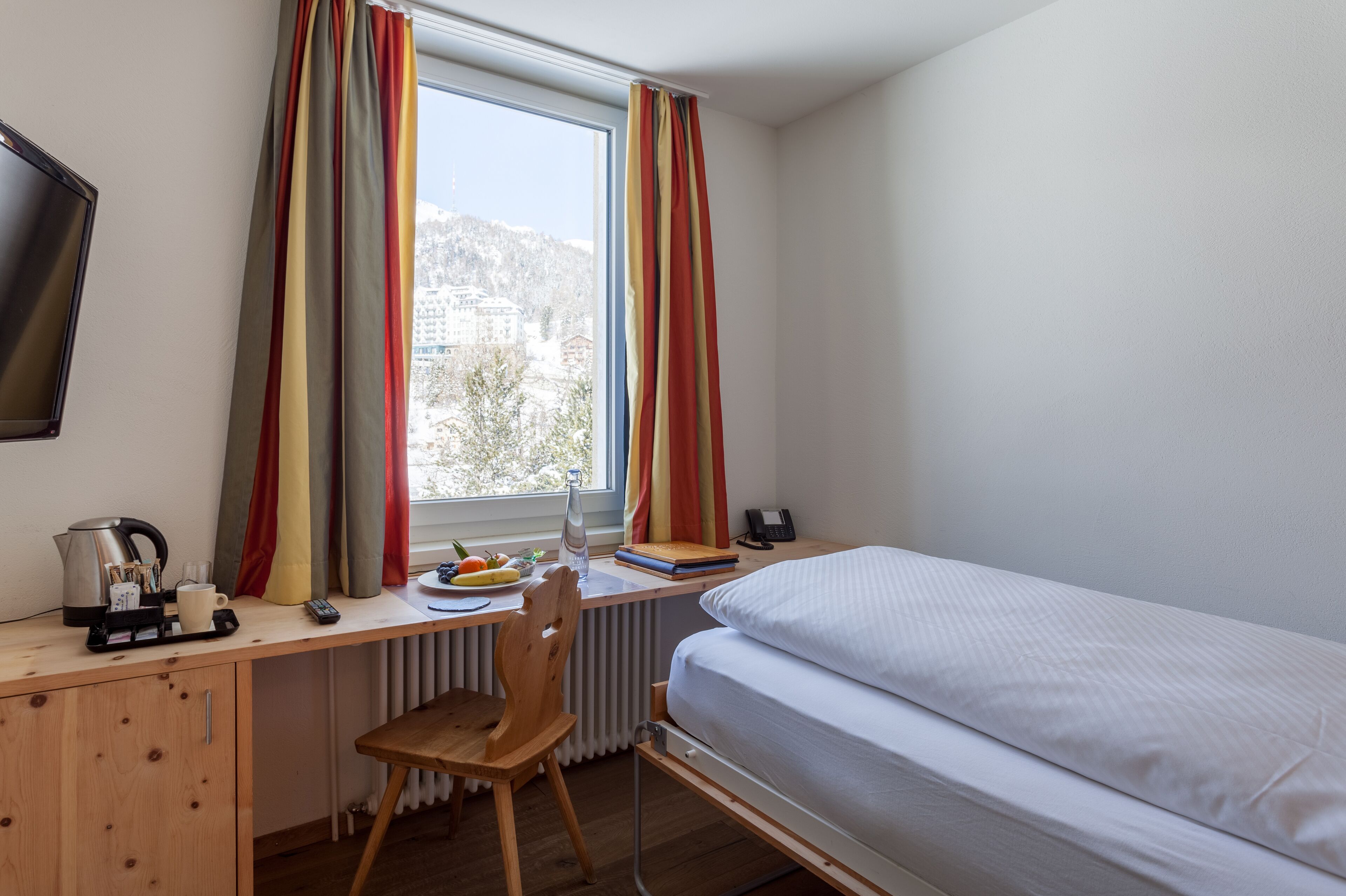 Una habitación con cama, un escritorio con silla, un radiador, una ventana con cortinas y vistas a edificios.
