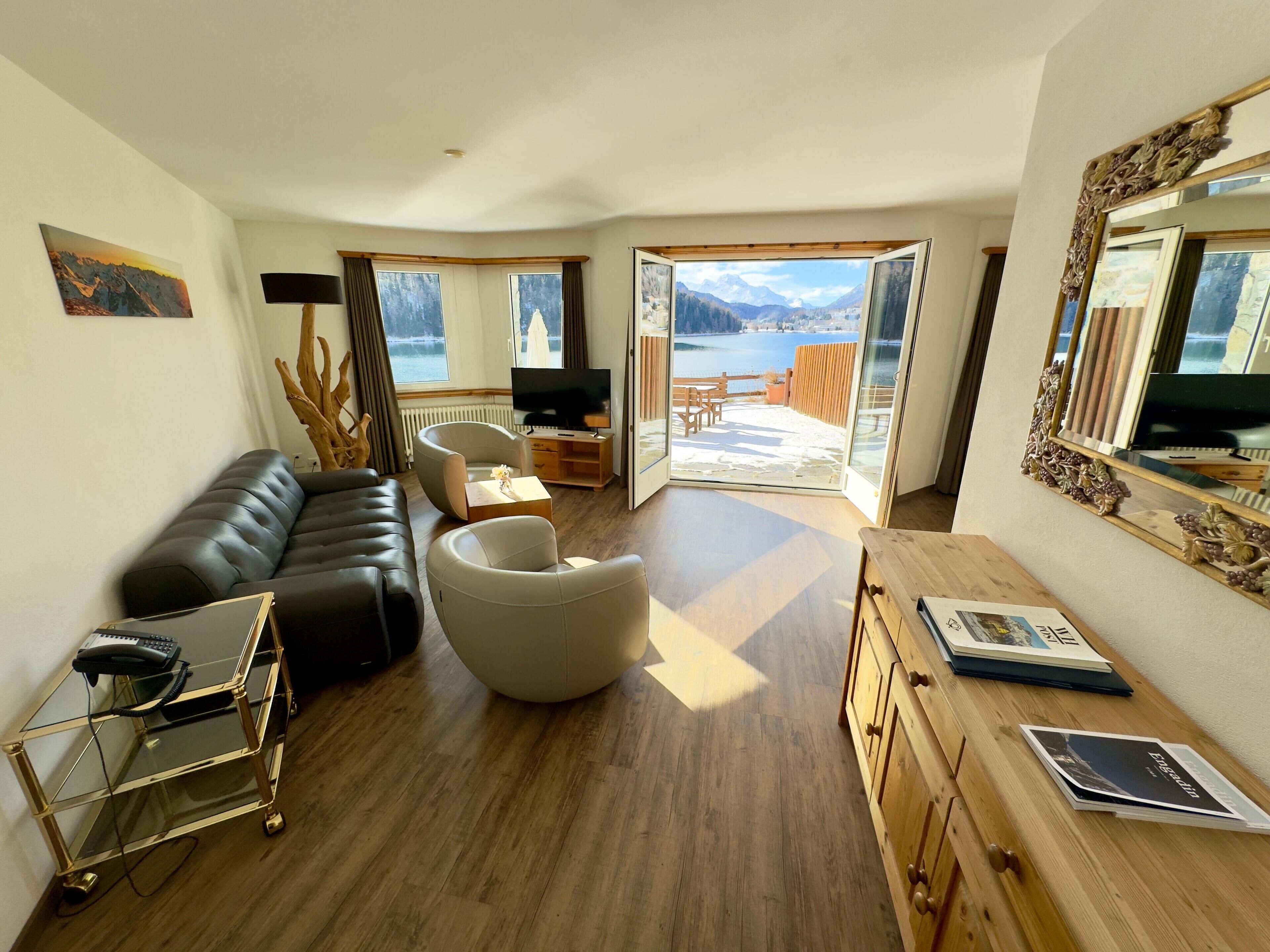 A modern living room with a leather sofa, a glass coffee table, and a television. There is a wooden cabinet with books and a mirror. The room has large windows with a view of a lake and mountains.