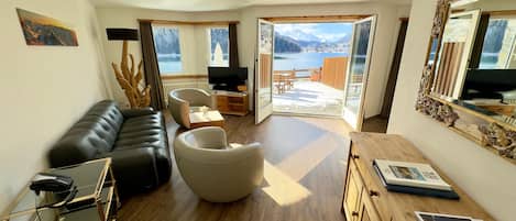 A modern living room with a leather sofa, a glass coffee table, and a television. There is a wooden cabinet with books and a mirror. The room has large windows with a view of a lake and mountains.
