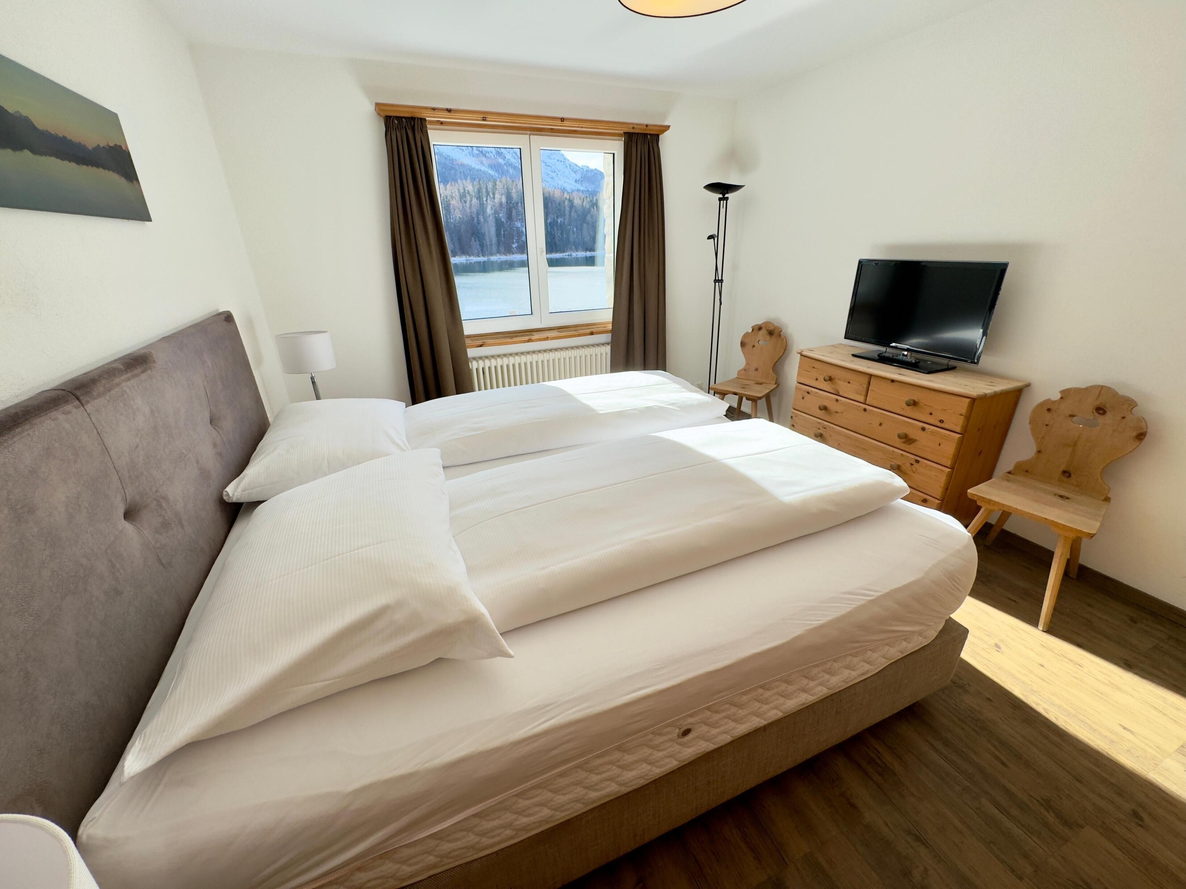 Ein Hotelzimmer mit einem großen Bett, einem Fernseher, einer Holzkommode und Blick auf Berge durch das Fenster.