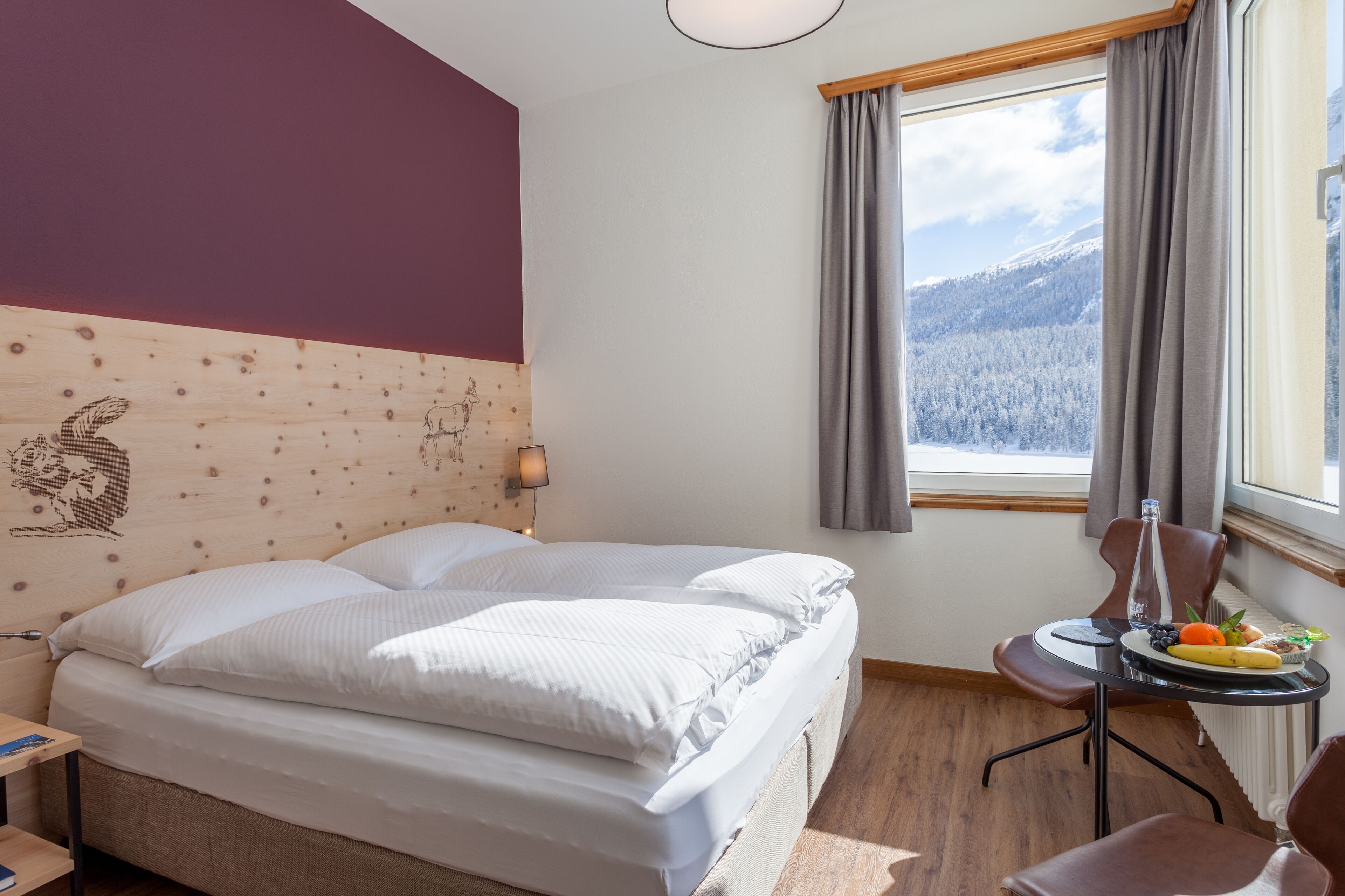 A bedroom with a wooden headboard, a bed with white bedding, a small table with fruits and a bottle, and a window with a view of snow-covered mountains.