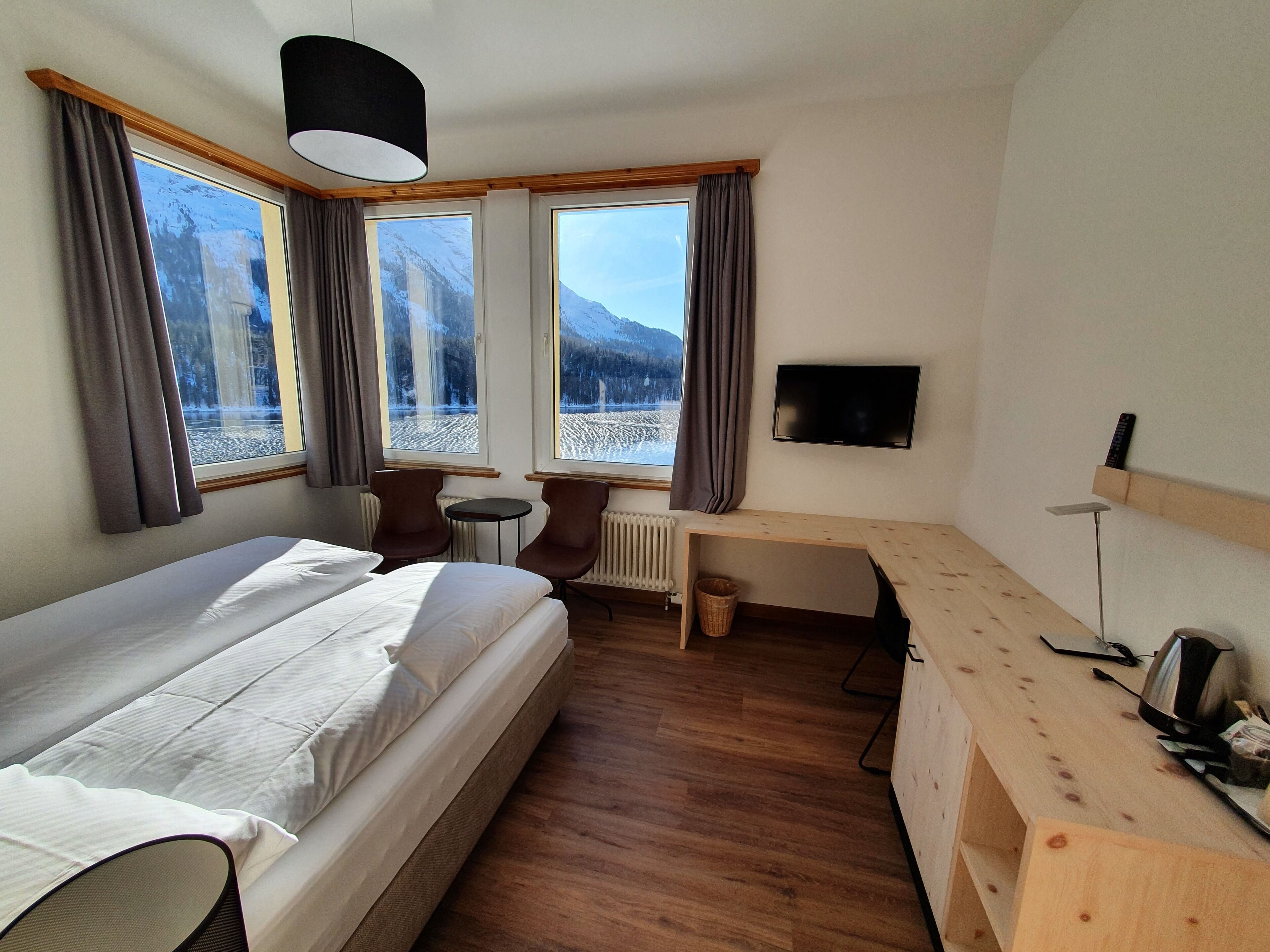 Habitación de hotel con cama, dos sillones, escritorio, televisor y ventanas amplias con vista a la montaña.