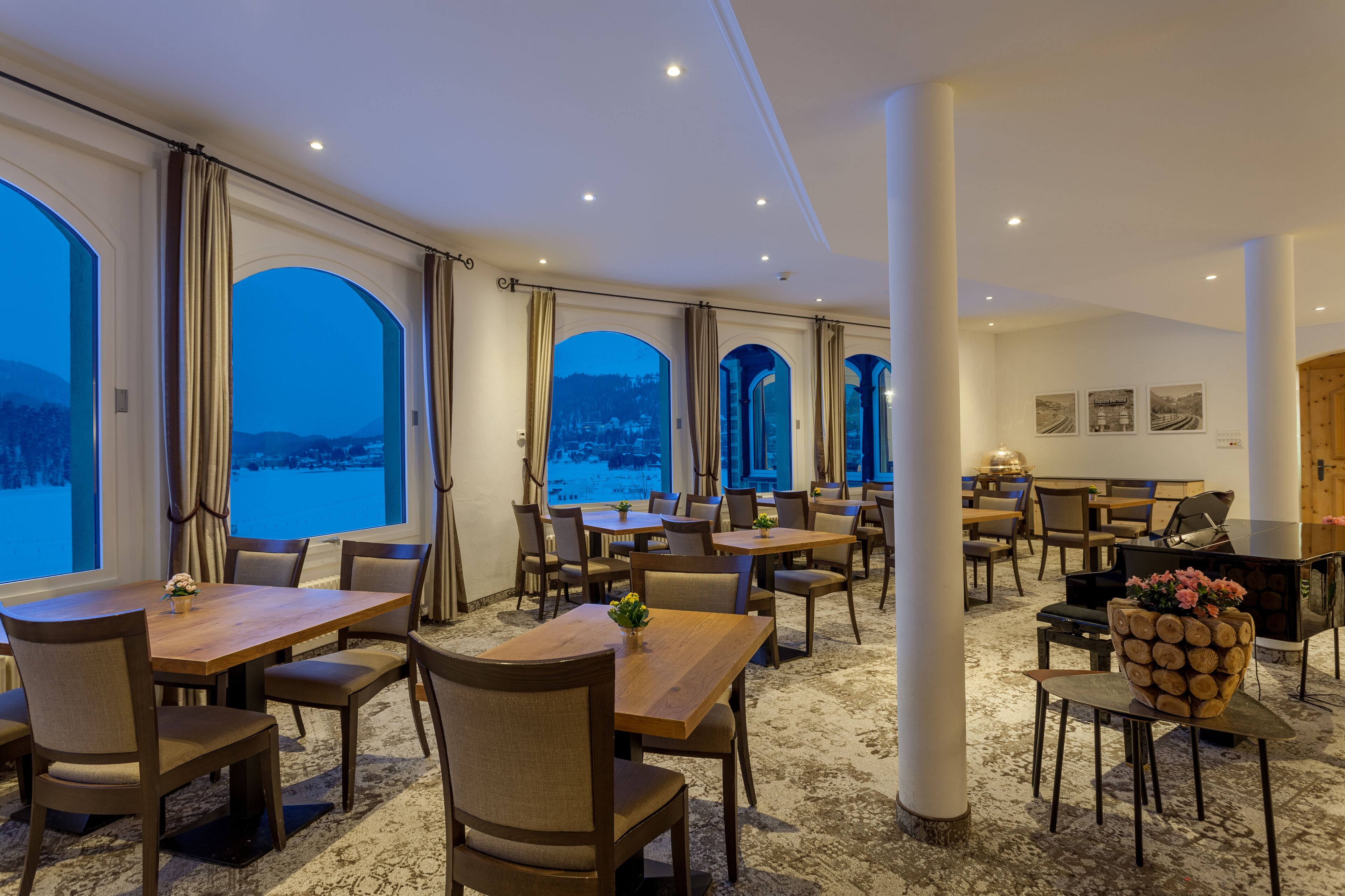 A hotel dining area with large windows, multiple tables, and chairs, and a piano in the corner.
