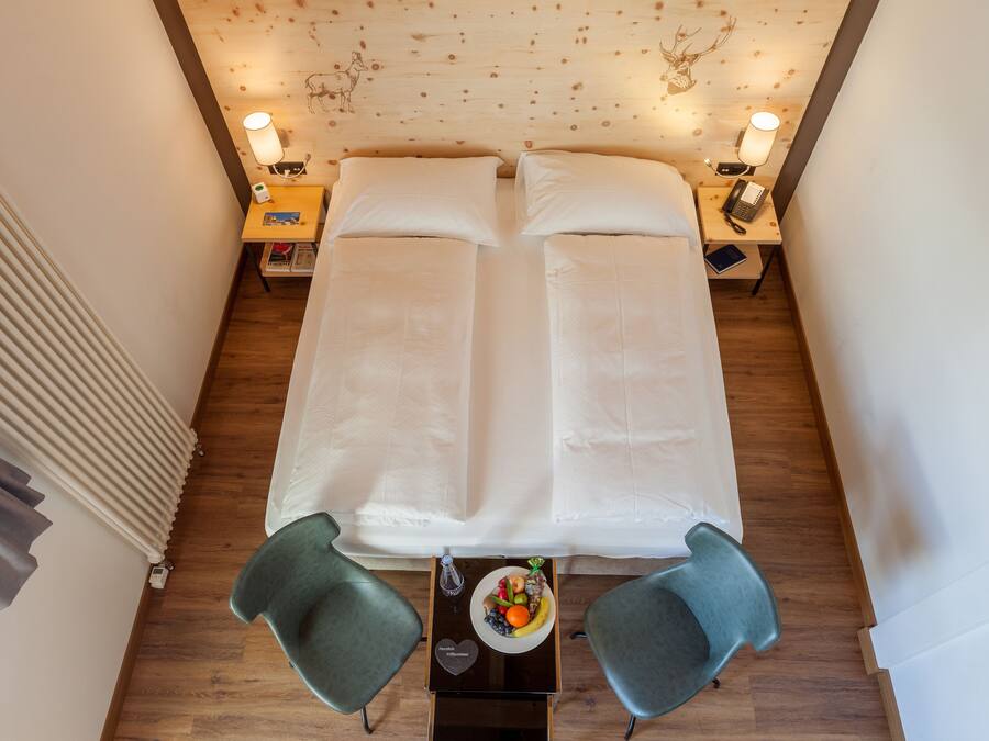 A double bed with white linens, two bedside tables with lamps, a small table with fruit and a heart-shaped object, and two green chairs.
