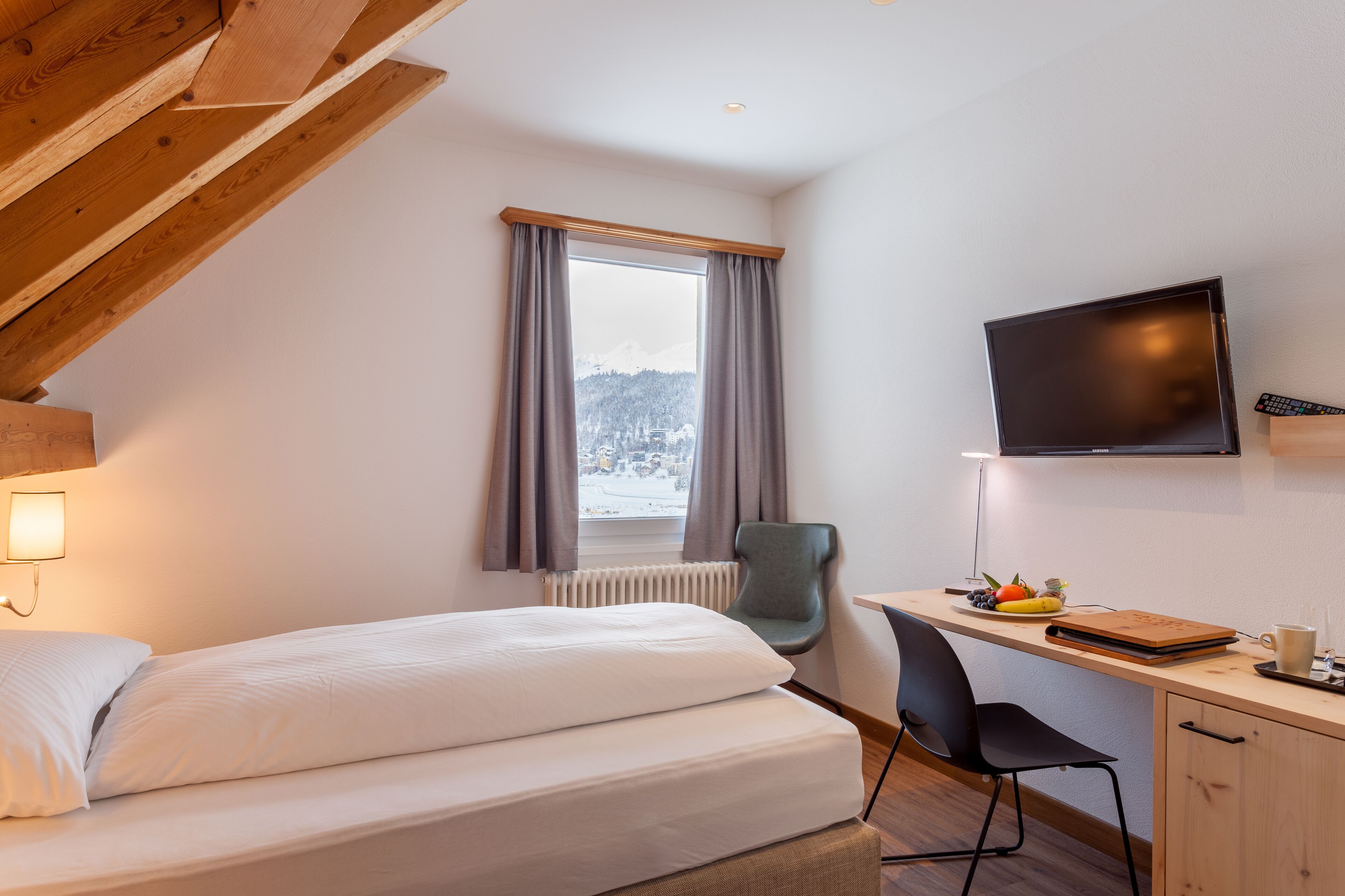 A bedroom with a bed, a desk, a chair, a TV, and a window with curtains.