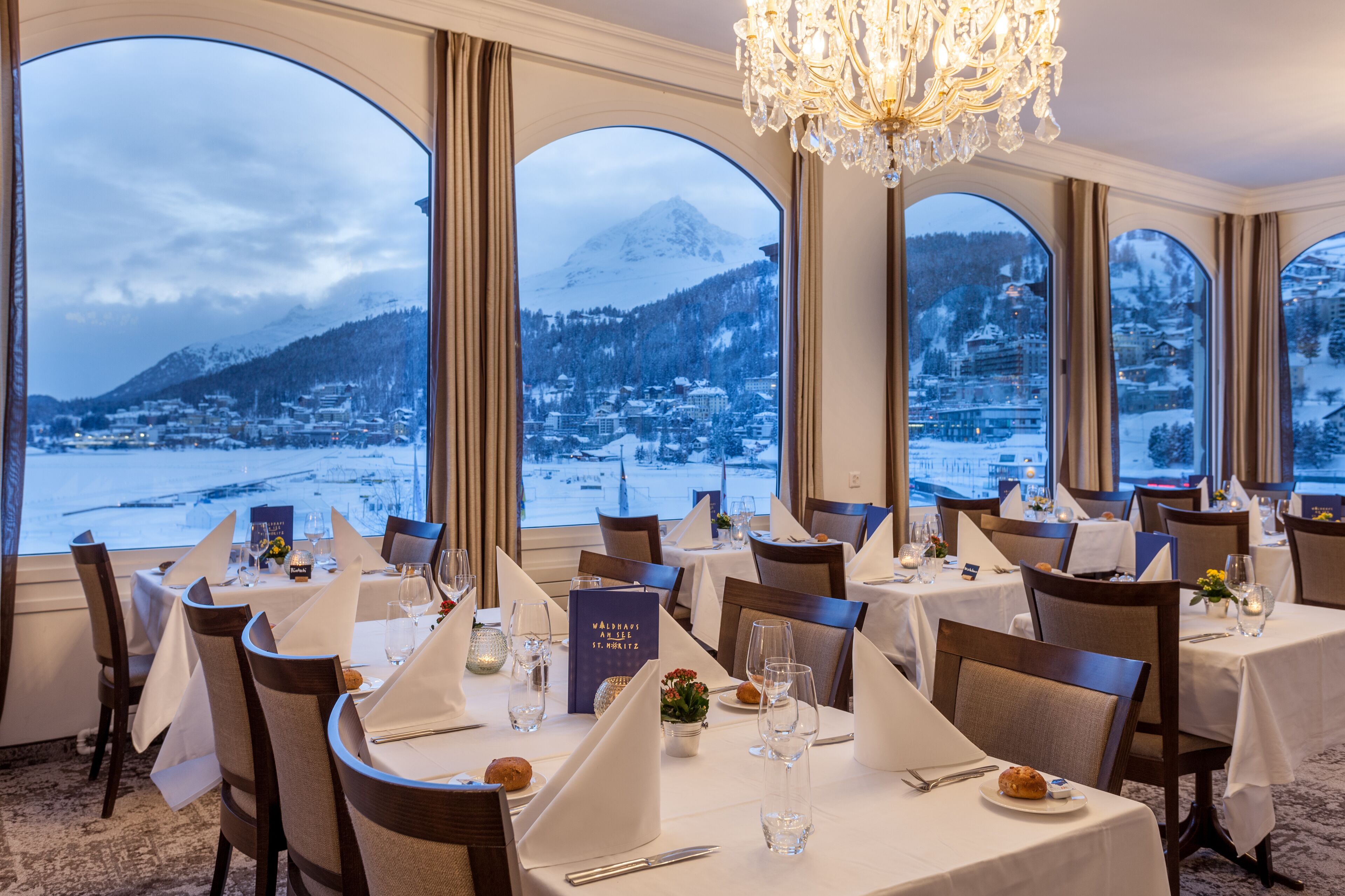 A restaurant with tables set for dining, large windows offering a view of a snowy mountain and town.