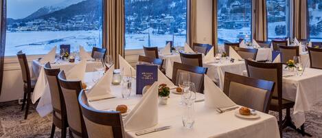A restaurant with tables set for dining, large windows offering a view of a snowy mountain and town.