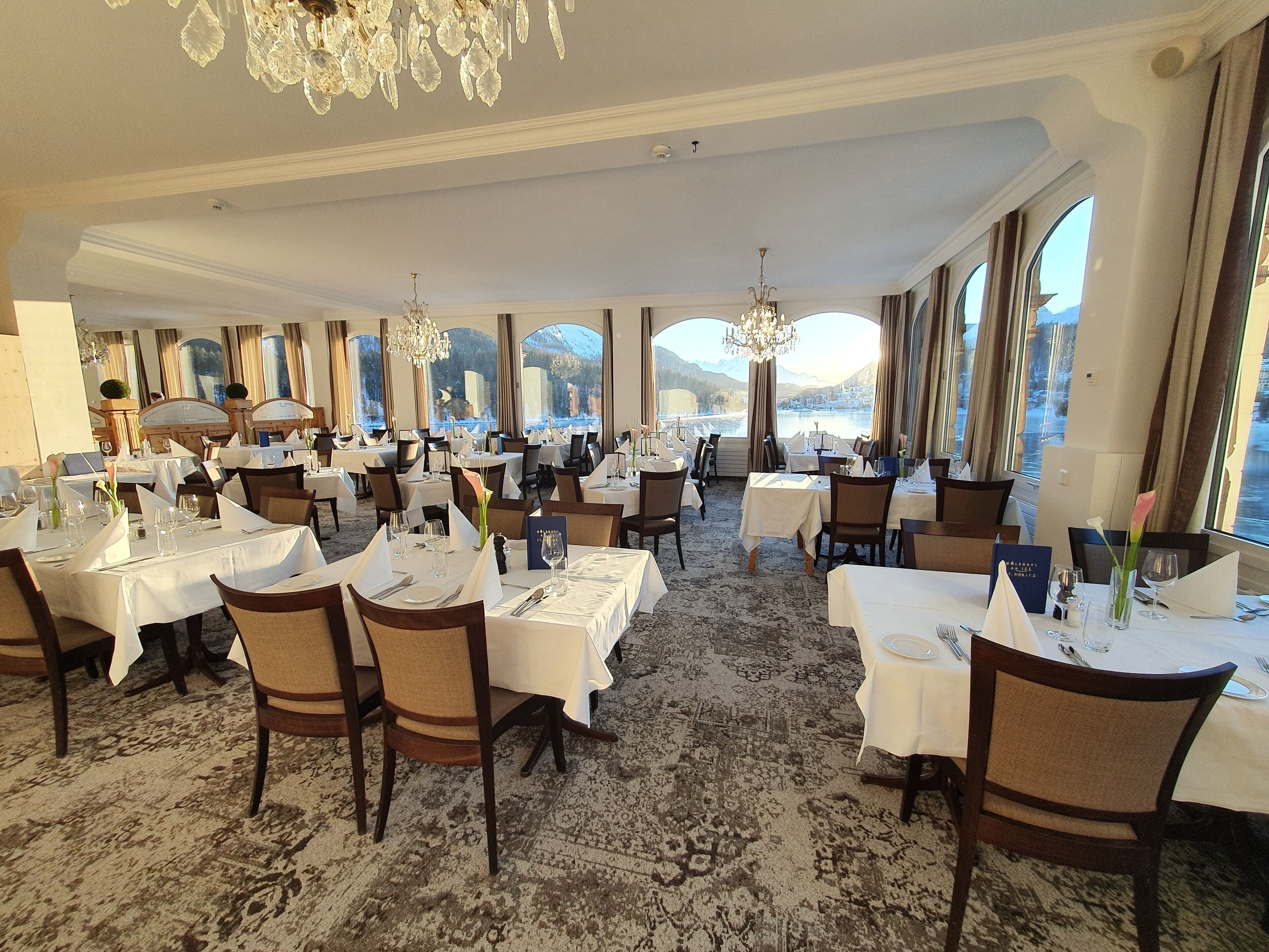 A spacious dining area with multiple tables set for dining, large windows offering a scenic view, and elegant chandeliers.
