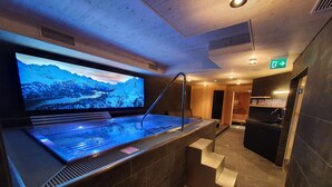 Zona de sauna con una pantalla grande que muestra un paisaje de montaña, un jacuzzi y una pequeña cocina con lavabo y encimera.